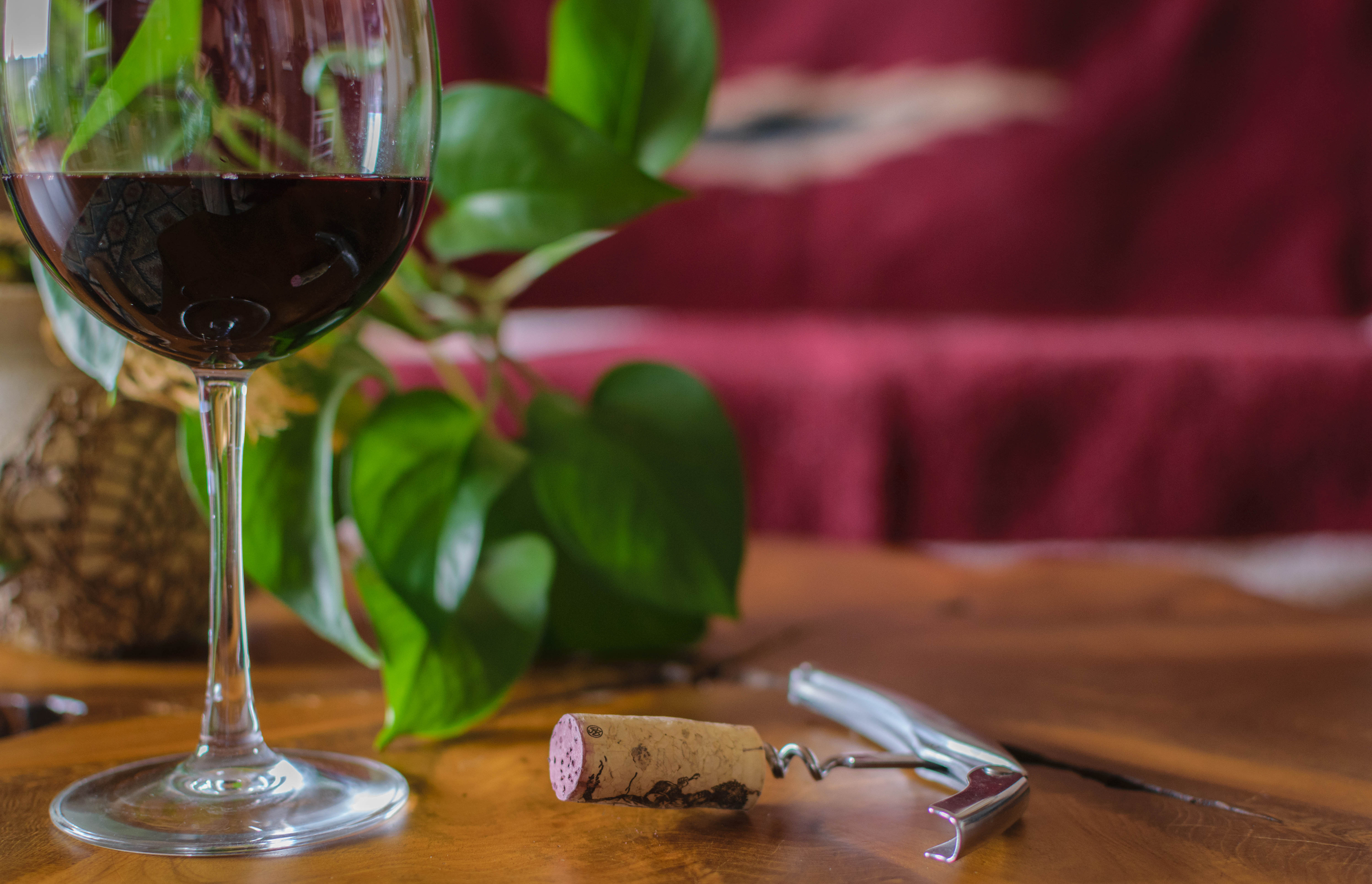 A glass of red wine sits on a wooden table with a cork, corkscrew, and green plant in the background.